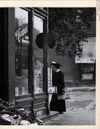 صورة رأسية بالأسود والأبيض تظهر امرأة تحمل سلة أثناء وقوفها أمام مدخل صيدلية WC Kalis في 4878 Easton Avenue بواسطة تشارلز كليمنت هولت